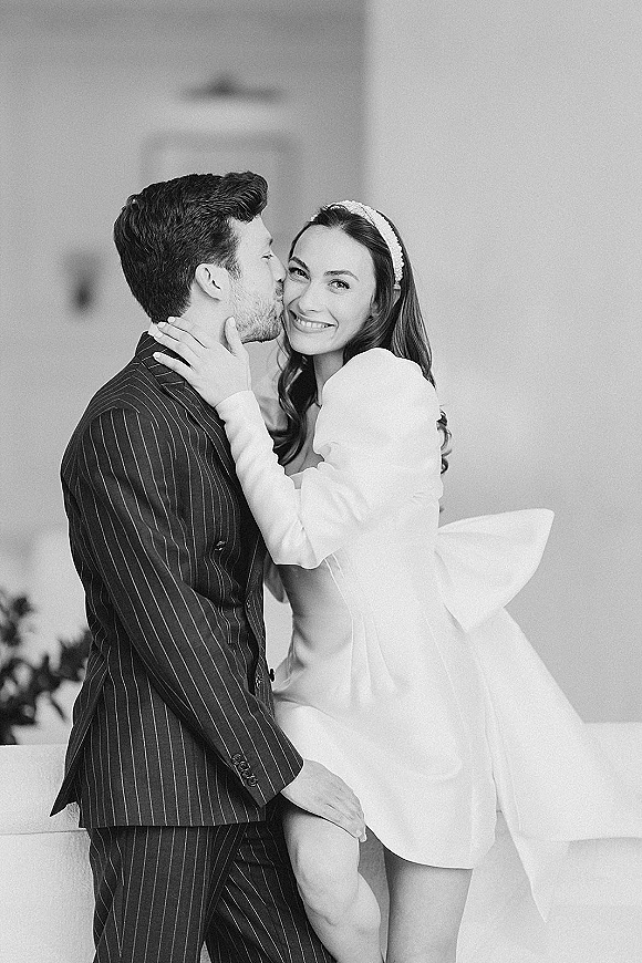 Wedding couple portrait of groom kissing bride’s cheek as she smiles, wearing a white mini dress with oversized bow in a simple indoor room