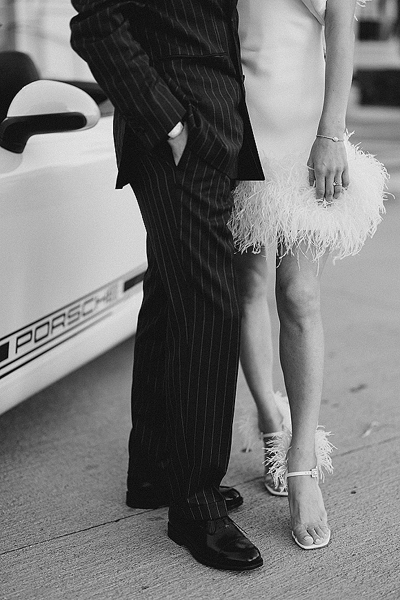 Couple portrait in a black and white wedding photo, bride in satin mini dress leaning by a luxury car on street pavement