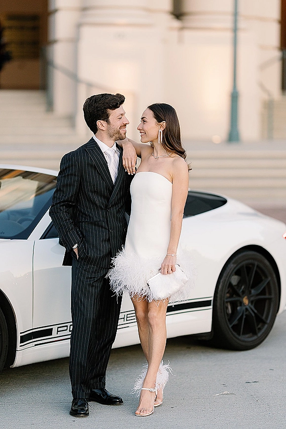 Couple portrait of bride and groom by car, her strapless feather-trim mini dress and his pinstripe suit on stone steps by a sports car