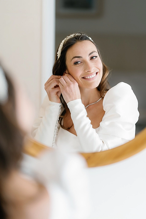 Bride getting ready, bride putting on earrings in a mirror reflection, wearing a wedding dress with pearl headband, necklace and earrings indoors