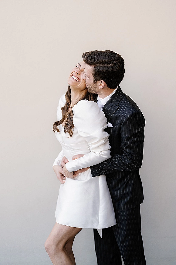 Couple portrait of bride in a short wedding dress laughing as groom in a pinstripe suit hugs her from behind against a neutral studio backdrop