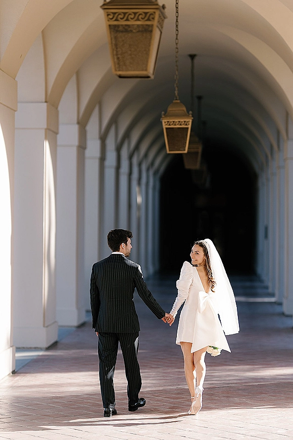 Couple portrait of bride and groom holding hands, bride in short wedding dress with veil and bouquet, walking through arched colonnade lanterns