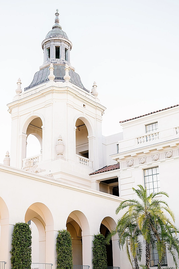 Wedding venue exterior with white stucco facade, arched colonnade, dome tower and red tile roof, framed by hedges and a palm tree under bright sky
