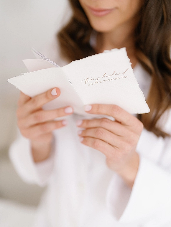 Wedding letter held by a bride in a white robe, letter to my husband on deckle edge paper with vow book and bridal manicure in soft light