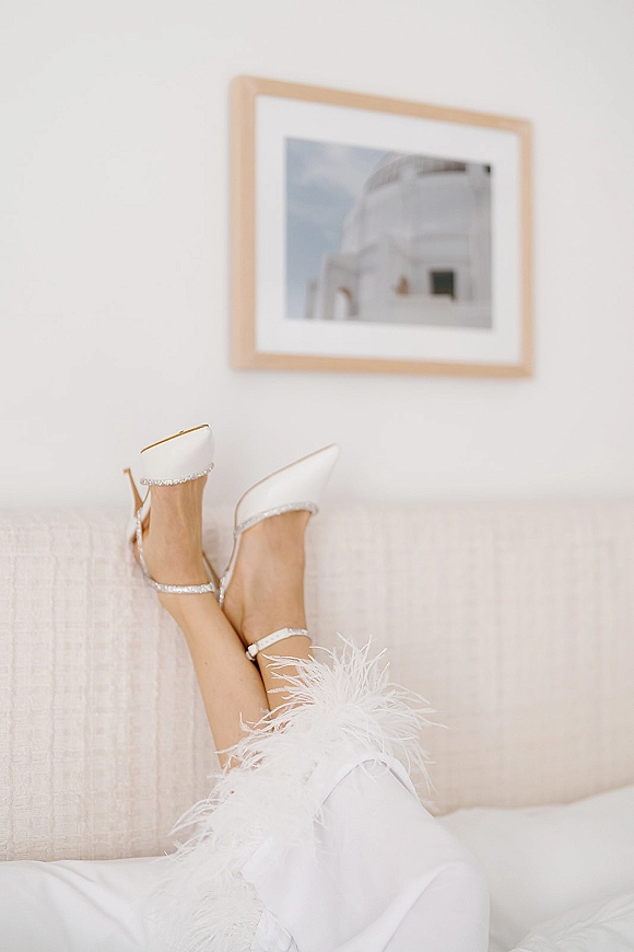 Bridal shoes, white wedding heels with rhinestone ankle straps on a white bed beside a feather-trim robe in a neutral getting-ready room