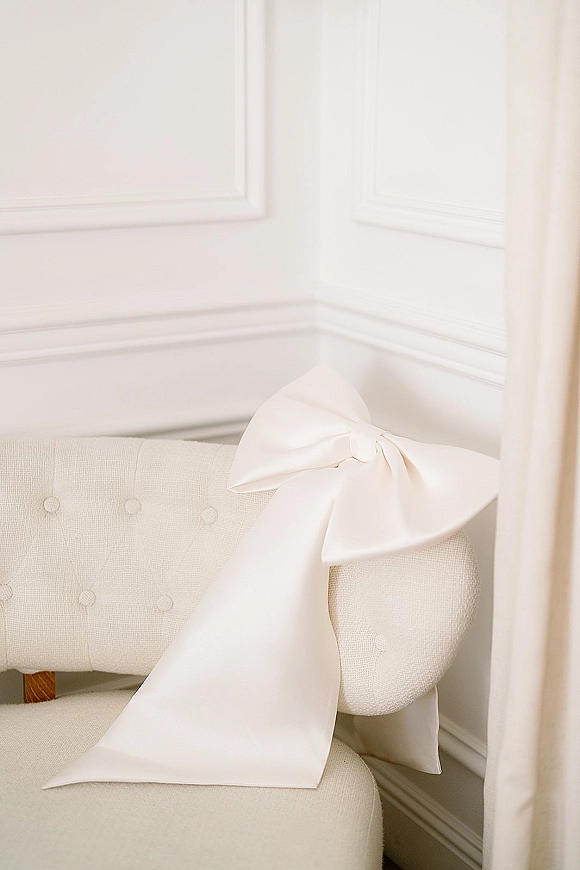 Bridal hair bow with an oversized satin bow draped in long tails on an upholstered chair beside a white paneled wall and sheer curtain