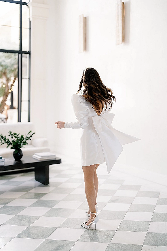 Bridal portrait of a bride in a short wedding dress with an oversized bow, twirling in a bright white room by a large window