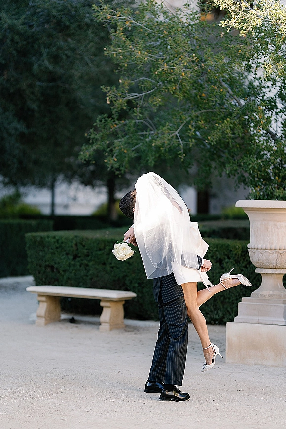 Wedding kiss portrait as groom lifts bride under her veil, bouquet in hand, leg kicked up on a gravel garden path by stone urns