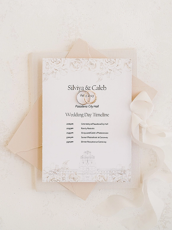 Wedding invitation suite with a wedding day timeline card, vellum ribbon, and gold rings arranged on a white textured surface flat lay