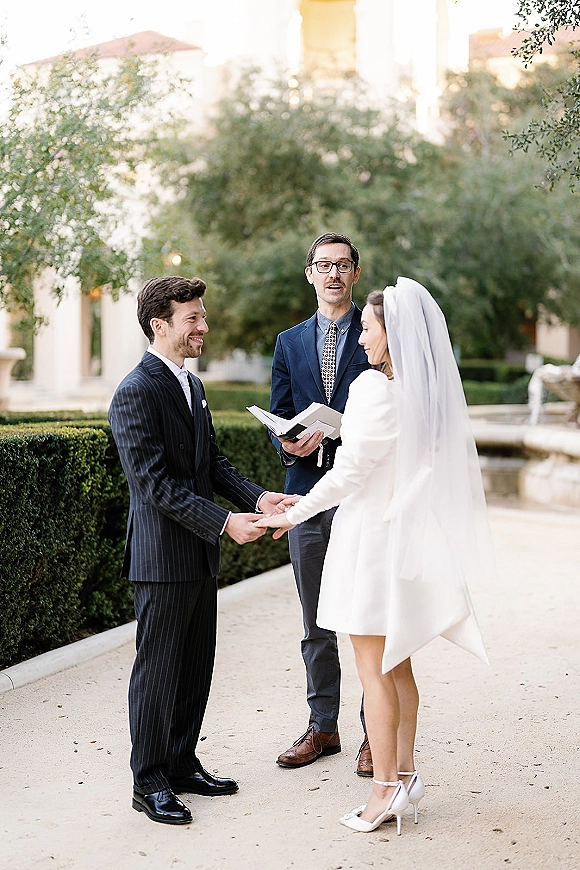 Wedding vows as bride and groom hold hands while officiant reads from a book in a garden courtyard with fountain backdrop