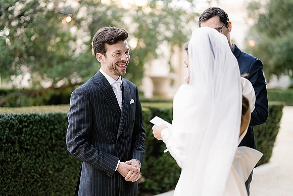 Wedding ceremony moment with outdoor wedding vows as bride in long veil reads from paper while groom in pinstripe suit smiles on a garden walkway