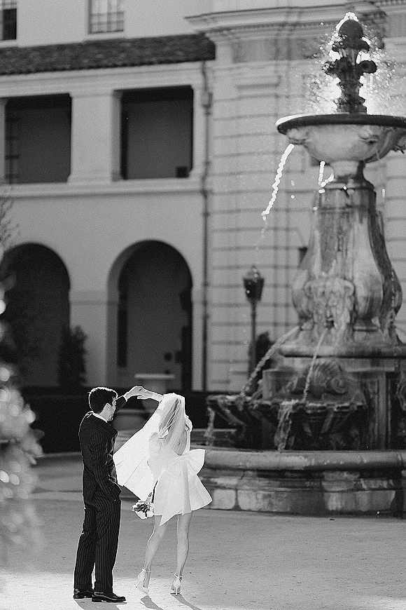 Couple portrait of bride twirling groom, veil flowing as she lifts a bouquet in a villa courtyard with fountain and arched doorways