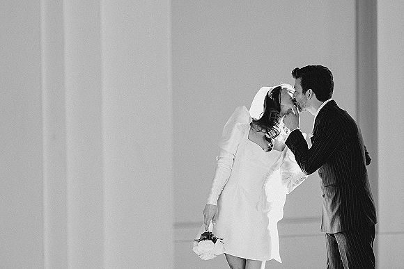 Wedding kiss portrait of bride and groom kissing, her veil and bouquet visible, groom in pinstripe suit against sunlit wall shadows