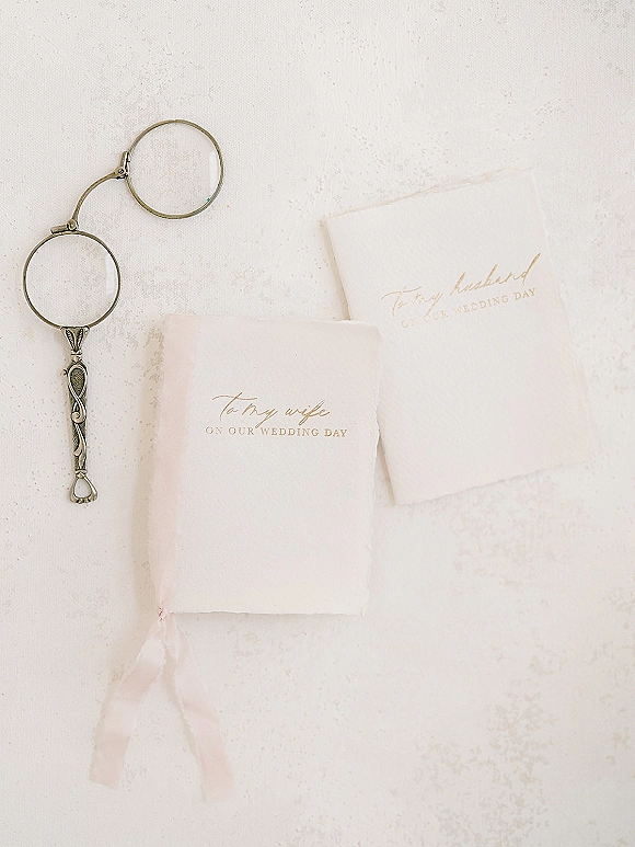 Wedding vow books with calligraphy covers tied with a blush ribbon, styled beside vintage lorgnette glasses on a white textured surface