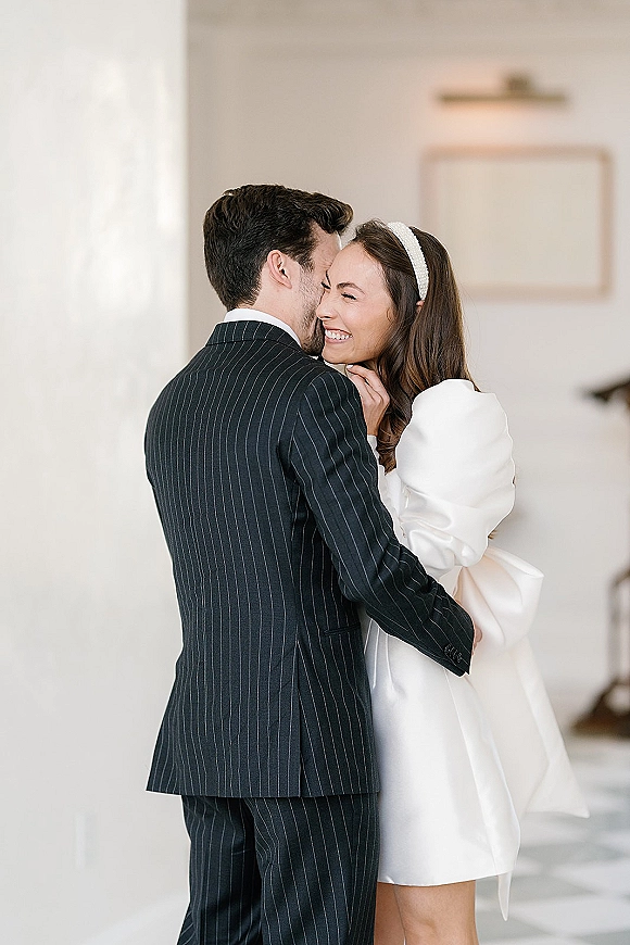 Couple portrait of bride and groom embrace, bride laughing in a puff-sleeve dress with pearl headband in a bright white room