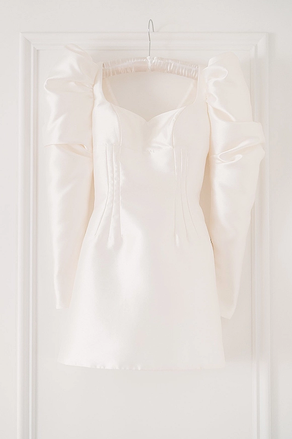 Wedding dress on a hanger, a short wedding dress in satin with puff sleeves and a sweetheart neckline against a white door and wall