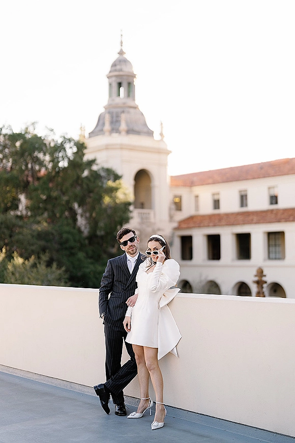 Couple portrait with wedding sunglasses, bride in a short dress and heels beside groom in suit on an outdoor terrace by arched windows