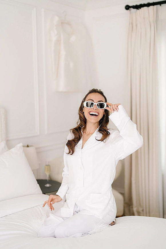 Bridal getting ready bride in pajamas laughs in sunglasses holding a champagne flute on a bed in a bright white bedroom, dress on hanger