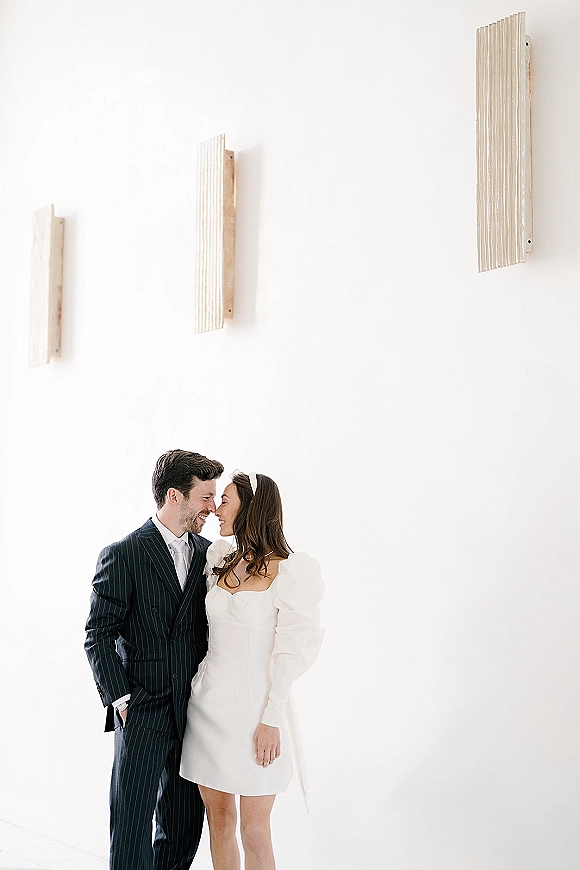 Couple portrait of bride in a short puff-sleeve wedding dress and headband nose-touching groom in a pinstripe suit by a white wall with sconces