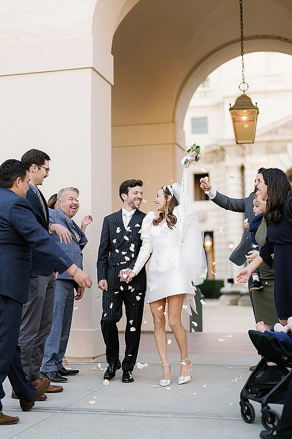 Wedding recessional with rose petal exit as bride in short dress lifts bouquet overhead, holding groom’s hand under an arched colonnade lanterns