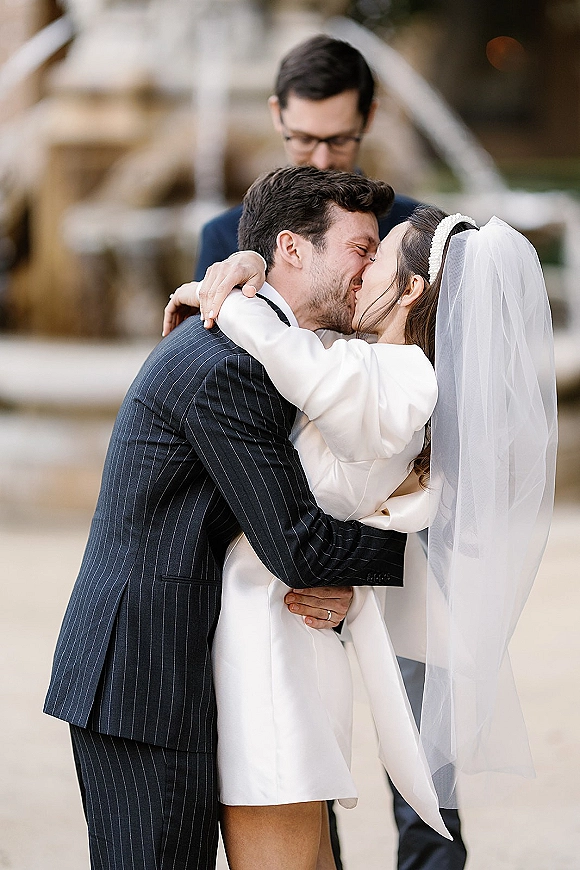 Wedding kiss as bride in short white dress and veil embraces groom in pinstripe suit with glasses on an outdoor walkway with greenery