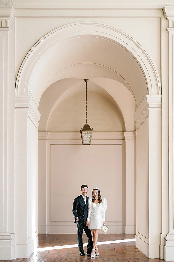 Couple portrait of bride and groom holding hands, her short wedding dress and bouquet framed by white columns under arched walkway lantern light