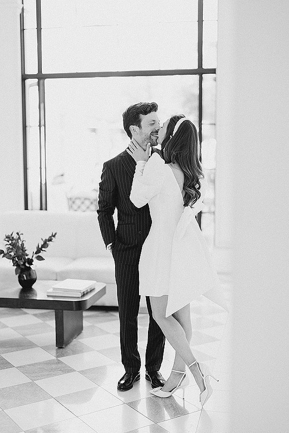 Wedding kiss portrait of bride kissing groom, her hand on his face, in a bright room with industrial windows and a white sofa
