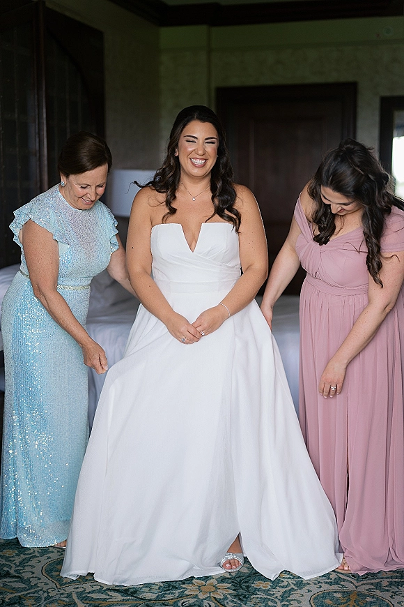 Bride getting ready in a strapless wedding dress as her mother and bridesmaid help with final touches in a softly lit bedroom