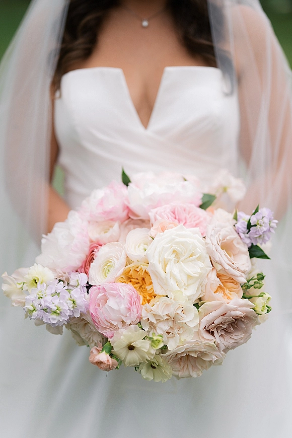 Bridal bouquet of pastel blooms and greenery held by a bride in a strapless dress and veil, with a softly blurred garden backdrop