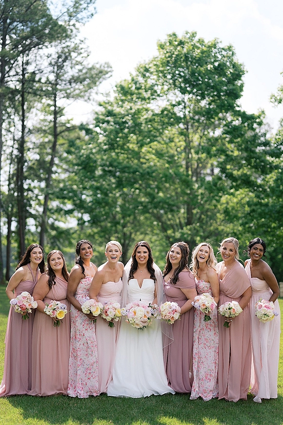Bridesmaids group photo with bride, holding pastel bouquets in mismatched blush and floral dresses on a green lawn with trees in daylight