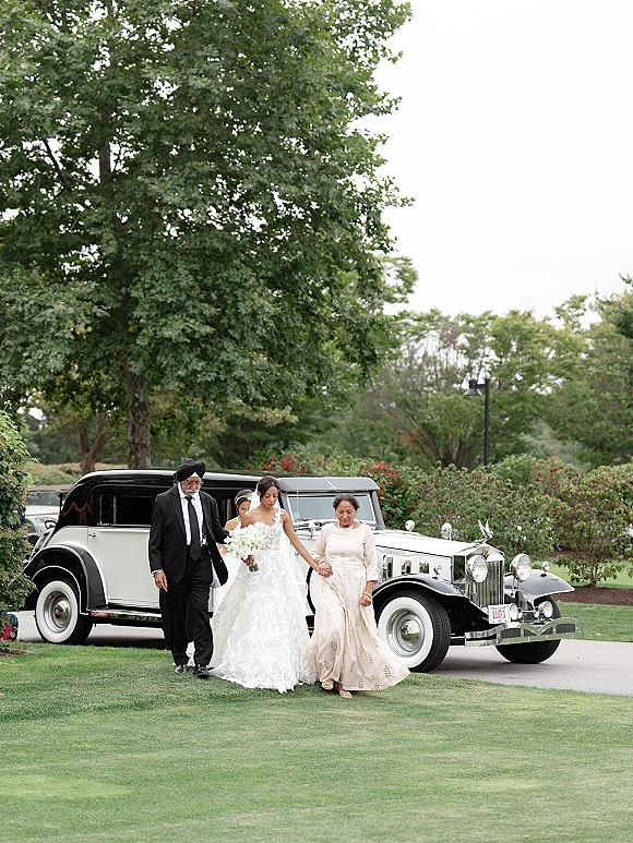Bride walking with parents, holding a white bouquet in a lace wedding dress and veil beside a vintage wedding car on a garden driveway