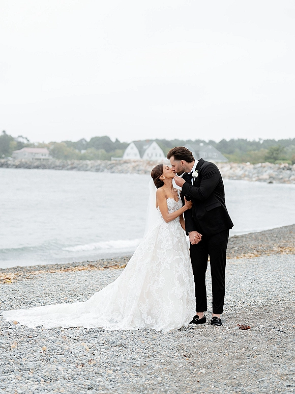 Wedding kiss portrait of bride and groom kissing on a rocky beach, her strapless lace gown and veil trailing as ocean waves roll in