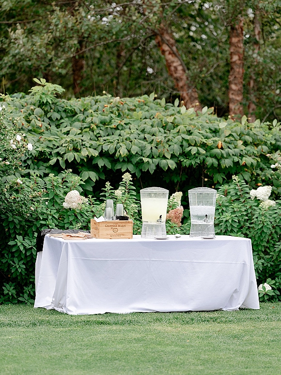 Wedding drink station with self serve dispensers and taps on a white tablecloth, clear cups, ice, and straws set by shrubs and hydrangeas