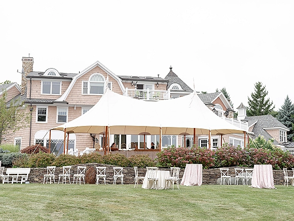 Outdoor wedding tent with white sailcloth canopy and wooden poles, set with cocktail tables, pink linens, and crossback chairs on a lawn