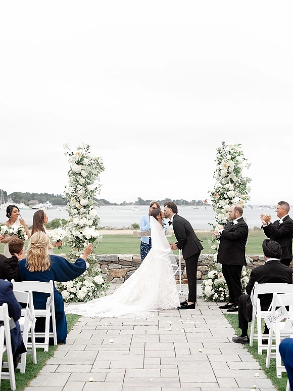 Ceremony kiss under a floral ceremony arch with white blooms, veil and train, on a waterfront lawn with boats under overcast sky