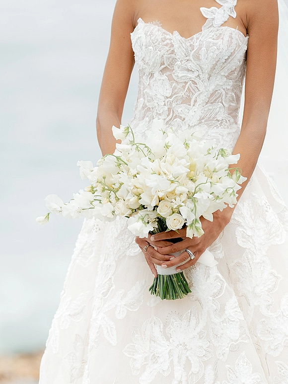 Bridal bouquet of white roses and sweet peas with ribbon wrap held against a lace halter wedding dress, manicure and ring visible