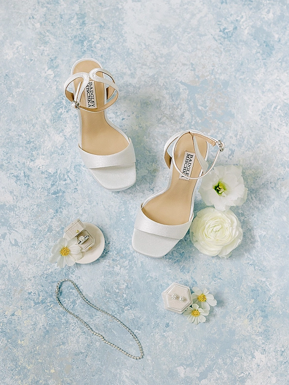 Bridal shoes flatlay of white wedding heels with ankle straps, rhinestone buckle, engagement ring and jewelry on a blue textured backdrop