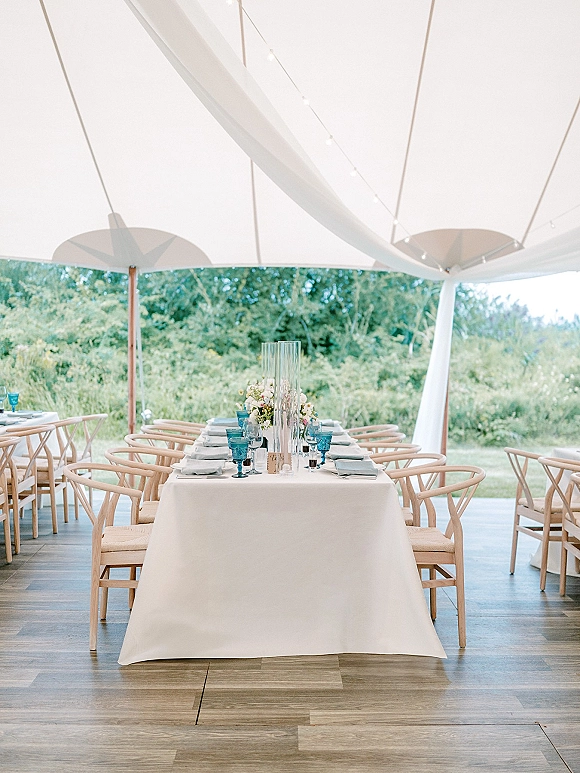 Reception tablescape in an outdoor tent reception with white tablecloth, blue goblets, greenery centerpiece, and string lights under draped ceiling