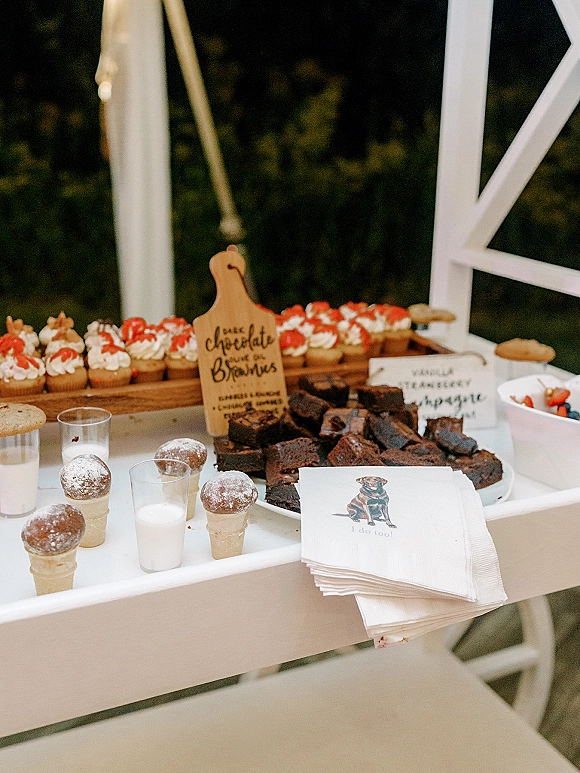 Wedding dessert table dessert bar on a white cart with brownies, cupcakes topped with berries, donut holes, and milk glasses by greenery