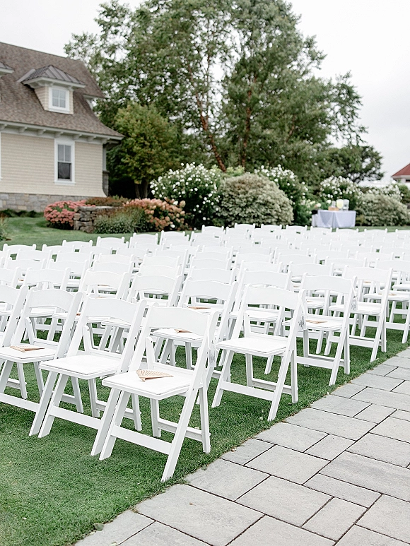 Outdoor ceremony seating with white folding chairs and programs beside a stone paver aisle on a lawn, with a drink table nearby
