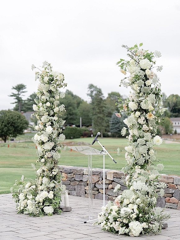 Wedding ceremony backdrop with floral ceremony pillars in white and blush blooms, greenery, and a clear acrylic podium before a stone wall