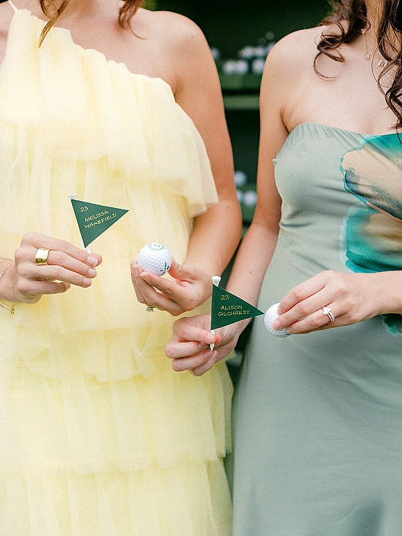 Bridesmaid portrait with bridesmaids holding props, showing yellow and mint dresses, jewelry, rings, and golf escort cards in greenery backdrop