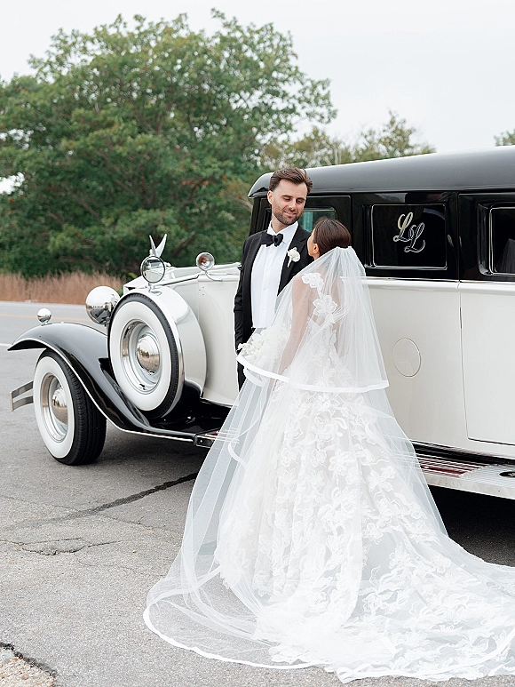 Couple portrait of bride and groom by vintage car, lace gown and long veil with tuxedo, standing on a tree-lined road under overcast sky