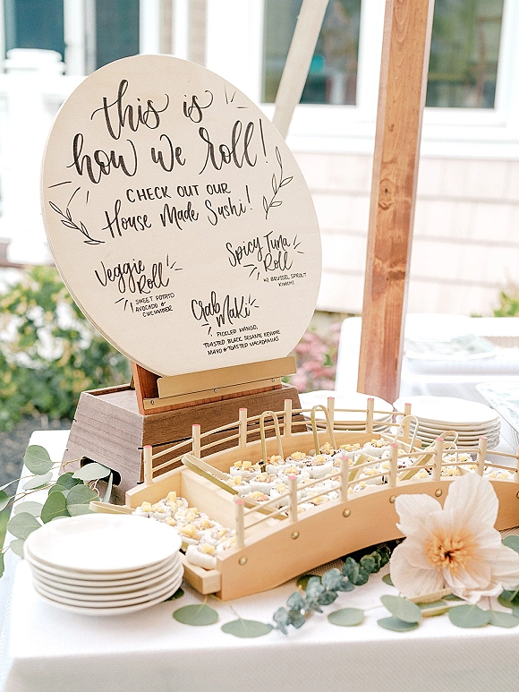Wedding sushi bar display with sushi rolls and small plates, a handwritten menu sign on wooden stand, and greenery garland on a white tablecloth