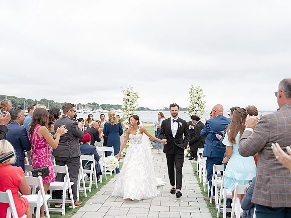 Wedding recessional as bride and groom walk down the aisle holding hands, veil flowing, guests clapping by a waterfront with boats under cloudy sky