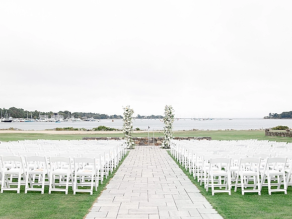 Ceremony setup with white folding chairs lining a stone aisle, floral pillars and clear acrylic podium overlooking a marina with sailboats