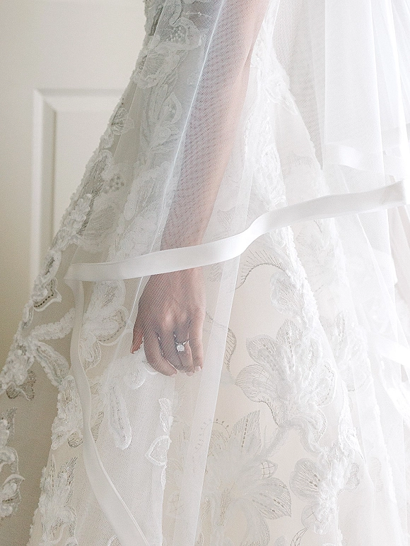Wedding dress detail with lace wedding dress appliqué and a bridal hand showing an engagement ring, set against a white wall in soft light