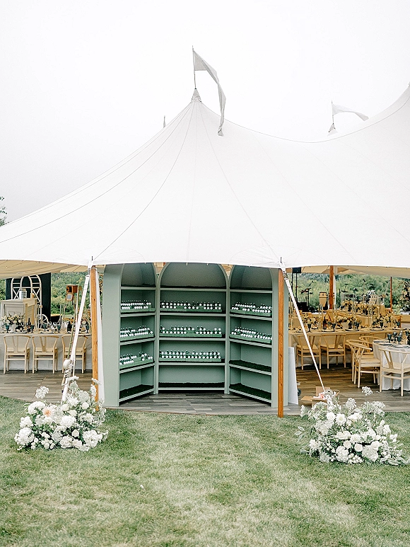 Wedding reception tent with sailcloth canopy, wooden poles, and string lights over round tables with white linens on a grassy lawn