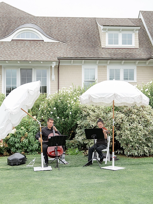 Wedding musicians in a string duo wedding play cello and violin from sheet music on a garden lawn beside white patio umbrellas and shrubs