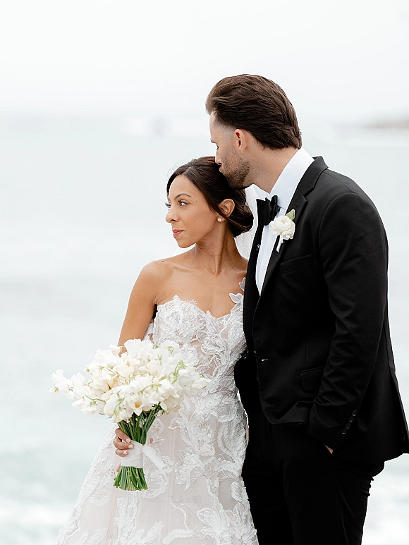 Couple portrait of groom kissing bride’s forehead as she holds a white bouquet, ocean horizon behind, strapless lace dress visible
